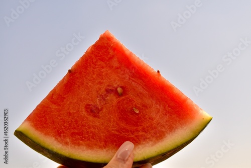 Hand holding a watermelon slice up to the sky on a bright sunny day. Summer, joy, and healthy nutrition.


