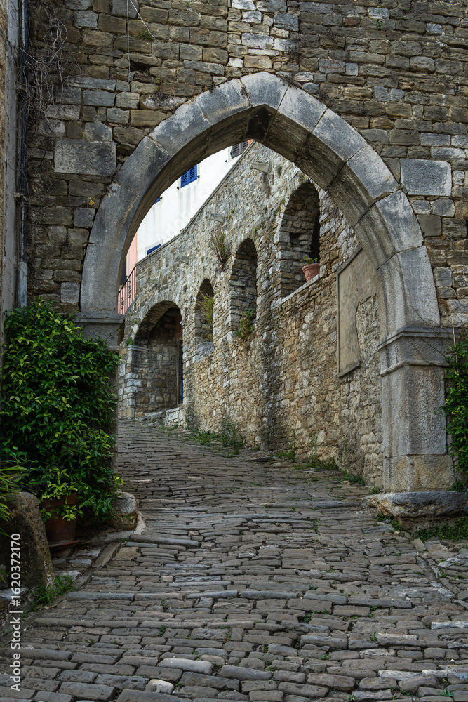 Obraz premium Historic stone street climbing uphill through arched passage in old medieval wall in Motovun, Istria, Croatia. Charming European townscape perfect for travel, history, architecture themes.