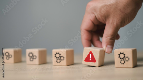 A hand picking a wooden block with an alert symbol among gear blocks, representing issue identification, risk management, and problem detection in process systems.