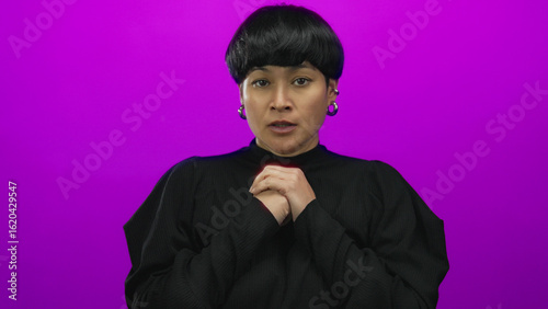 Woman with short hair and earrings appears anxious on an isolated pink background, hands clasped, projecting vivid emotion, in a minimalist setting showcasing expressive portraiture.