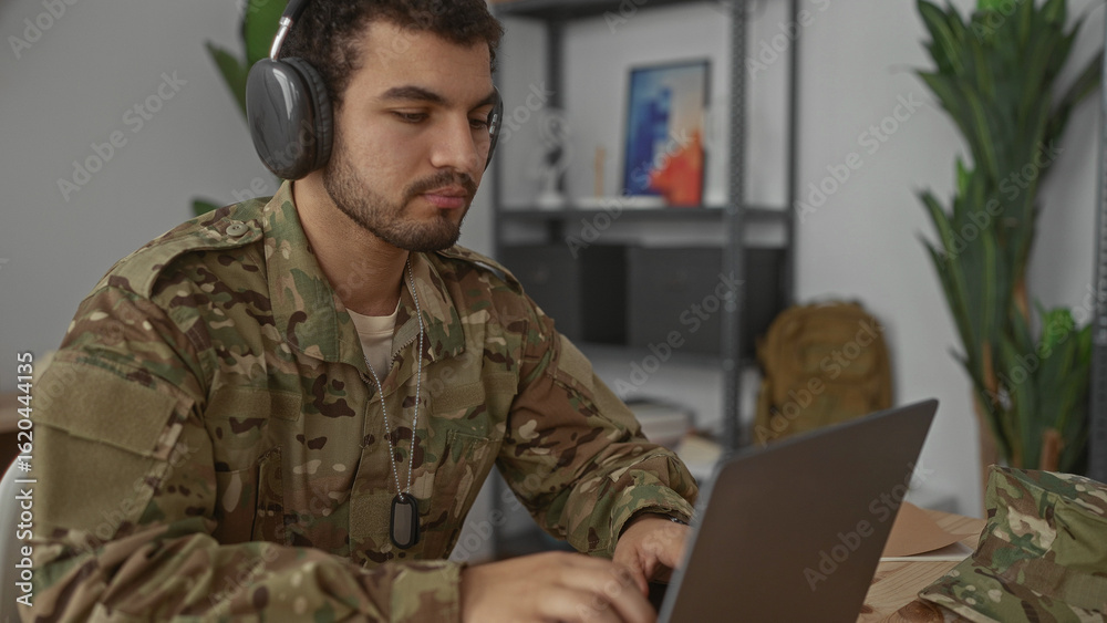 Fototapeta premium Man in camouflage uniform wearing headphones and dog tags typing on laptop in living room; focused discipline.