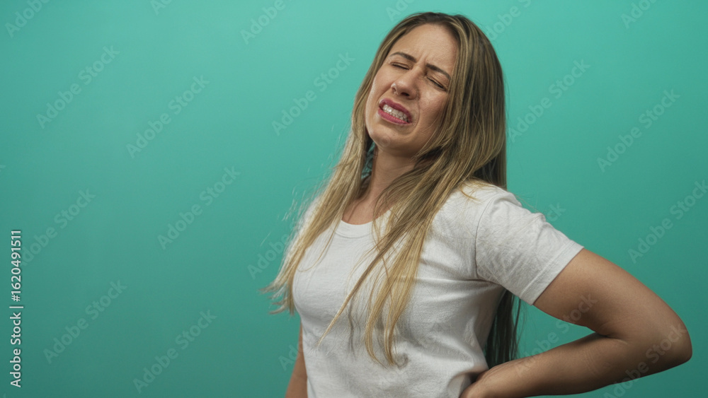 Fototapeta premium Woman clutching lower back in studio with teal background, leaning sideways and wincing in pain; discomfort tension stress.