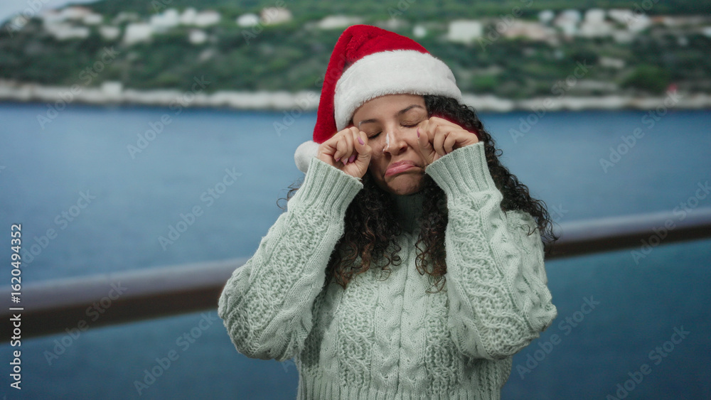 Obraz premium Woman wearing santa hat by the beach with a sad expression, dressed in warm sweater, showcasing emotions with the sea in the background on a cloudy day.