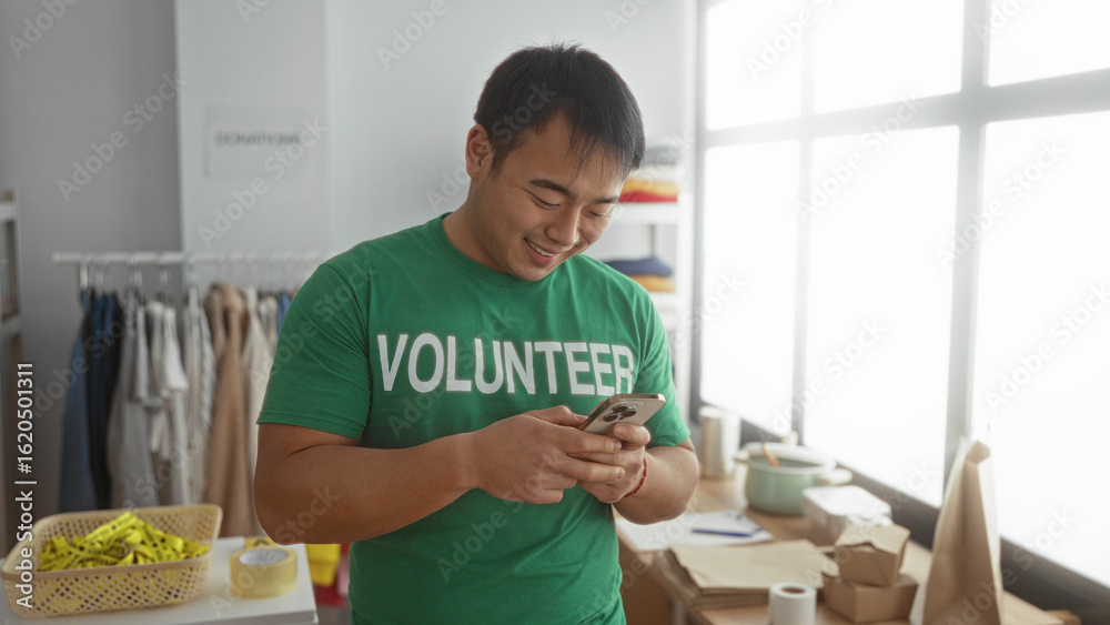 Fototapeta premium Man wearing green volunteer shirt using smartphone in donation center surrounded by boxes and clothes in bright room.