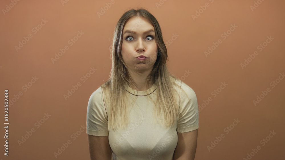Obraz premium Blonde woman playfully puffing cheeks isolated against brown wall background wearing casual shirt with exaggerated expression.
