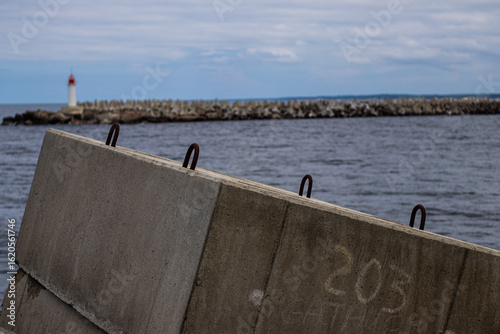 Schilderij op canvas Coastal scene with concrete barrier in foreground, distant lighthouse, and breakwater stretching into a moody sea under cloudy skies