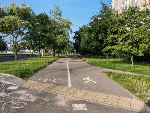 An asphalt sidewalk and bike lane with pavement markings run between buildings and the roadway of the street. Nearby grassy lawns, rowan, birch and other trees. Sunny summer weather and blue sky