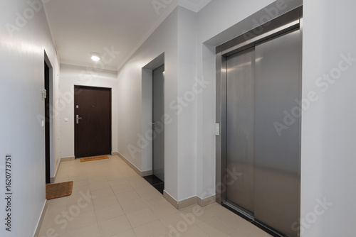 Interior of a modern apartment building hallway with a stainless steel elevator and entrance doors. This empty common corridor in a new residential complex is a perfect real estate background.