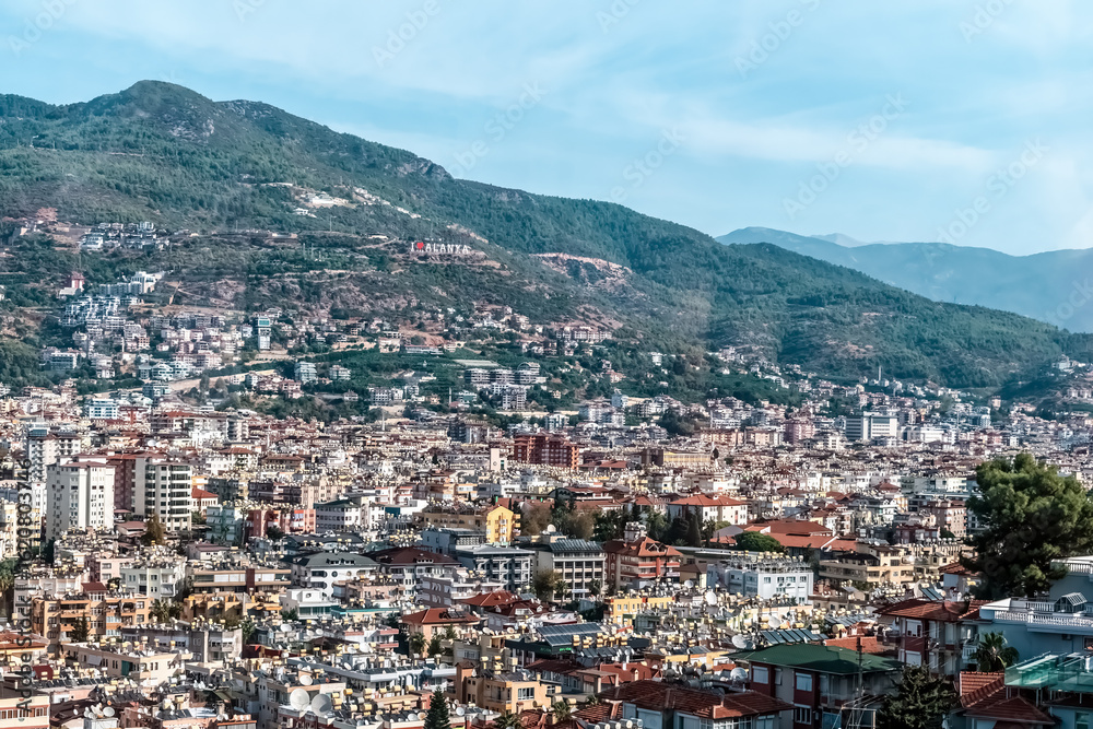 Naklejka premium Panoramic view of Alanya, Turkey, with the city's buildings stretching across the landscape and the Alanya sign visible on the hillside. The backdrop includes green mountains under a clear sky