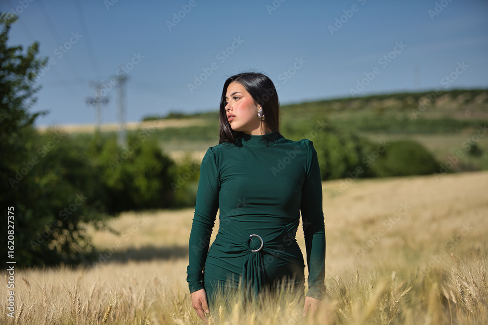 Obraz premium Young, pretty, brunette woman in a nice dark green dress, walking while watching the harvest of a ripe wheat field. Concept beauty, fashion, trendy, youth.