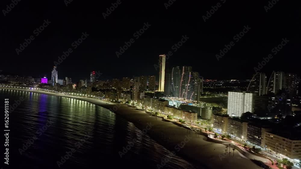 Night aerial shot of Benidorm, Spain, showing illuminated beachfront, high-rise buildings, and reflections on the sea in a vibrant urban coastline atmosphere. Night aerial shot of Benidorm, Spain, showing illuminated beachfront, high-rise buildings, and reflections on the sea in a vibrant urban coastline atmosphere.