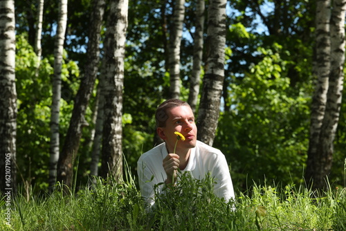 Young man with yellow dandelion in hand lies in the grass in a birch forest.