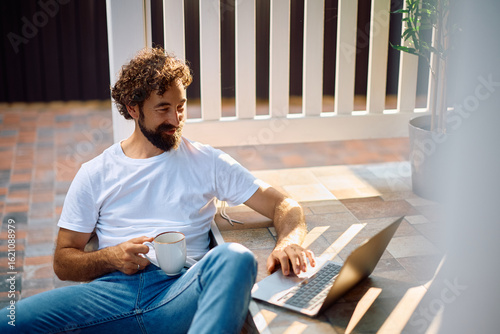 Fotografie Smiling man with cup of coffee surfing the net on laptop on porch