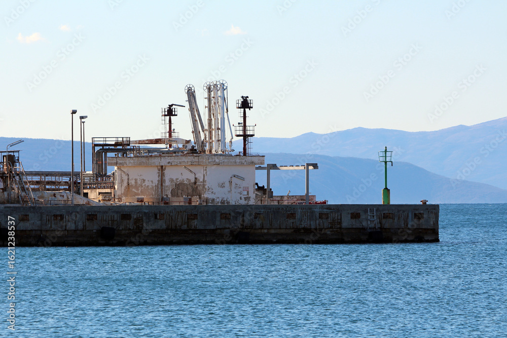 Fototapeta premium An aging dockside marine terminal features rusted equipment and industrial structures set along blue water, framed by distant mountains under a pale sky
