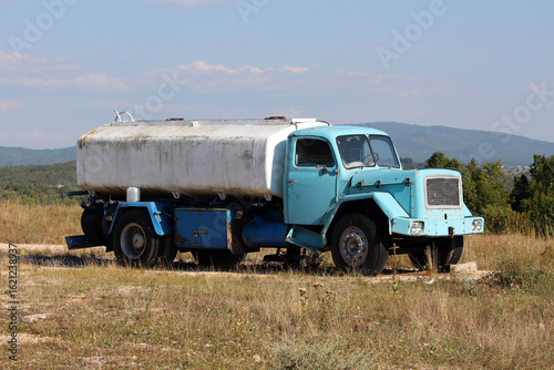 A vintage blue water tanker truck with a large white tank rests on uneven dry grass, its faded paint, rusty details, and missing rear wheels revealing years of service in a rural landscape