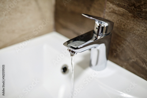 Modern chrome faucet with flowing water in a pristine white sink, surrounded by textured stone wall, showcasing contemporary bathroom design and functionality