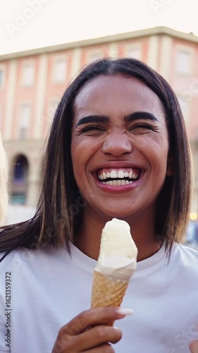 Vertical. Three young multiracial women enjoy ice cream cones together in a sunny Italian village square, smiling and chatting while sitting relaxed in a charming Mediterranean setting