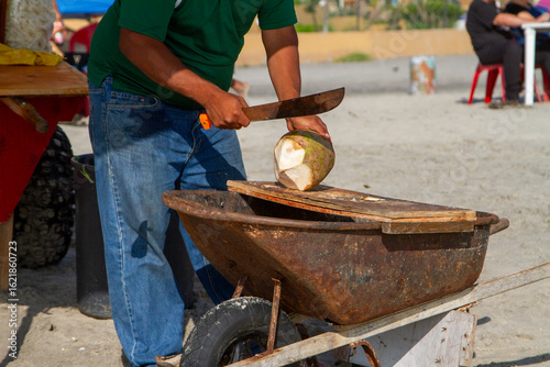 Close up of a man chopping a coconut with a macheti on a wheelbarrow on a beach