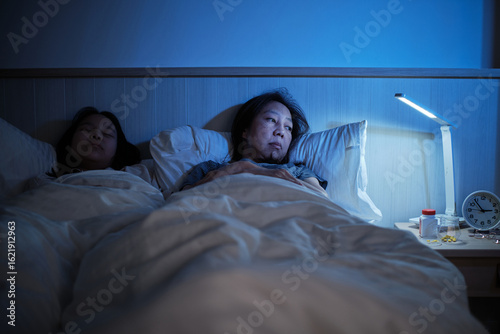 Photos Asian single mum is sleepless near her daughter, stressed, and worried awake at night because of drug side effects, insomnia illness in bed, mature and menopausal woman, family healthcare problem