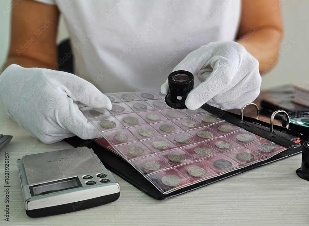 Fototapeta premium Coin collector examines rare coins with magnifying glass at a workstation concept