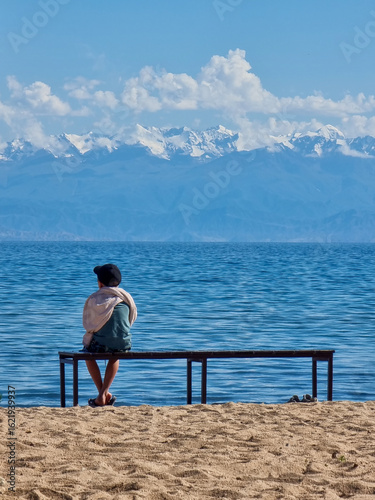 A boy sitting on a bench on the shore of a large blue lake against the background of mountains. Issyk-kul, Kyrgyzstan