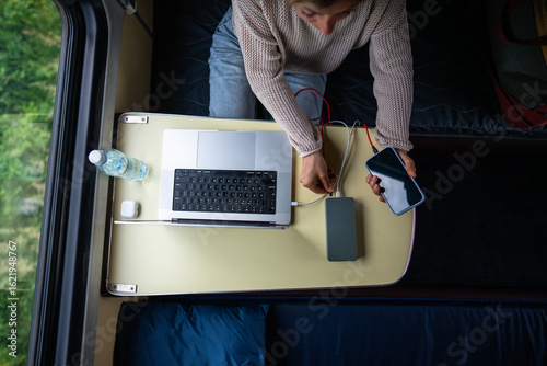 Tablou pe pânză Woman's hands plugging mobile phone and laptop into high-capacity power bank, charging batteries, sitting at table in train compartment, top view