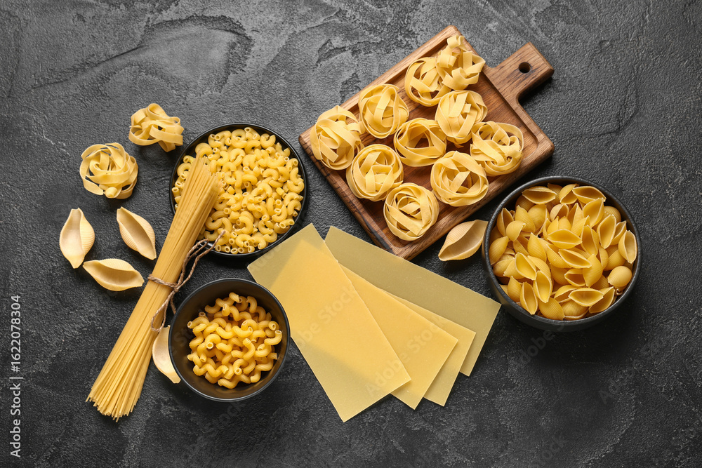 Fototapeta premium Cutting board and bowls with different types of raw pasta on black grunge background