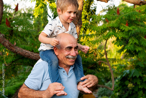 A smiling grandfather gives his grandson a shoulder ride, capturing a tender moment of joy, warmth, and playful connection in a vibrant outdoor garden setting