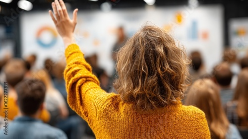 Woman in yellow sweater raises hand, front view of audience in meeting room, chart in background.