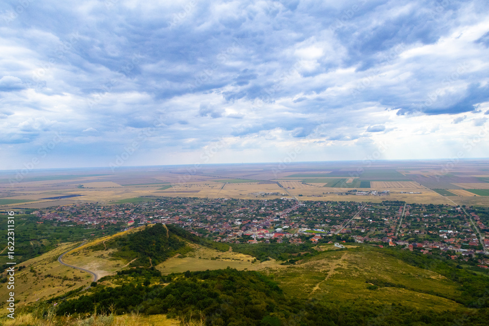 Obraz premium A small town nestles between forested hills and patchwork fields under a partly cloudy sky, blending rural life with nature in a serene, panoramic landscape.