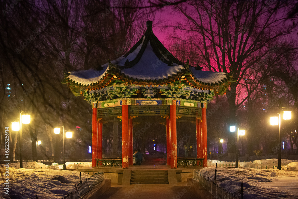 Fototapeta premium Colorful Chinese pavilion covered in snow, illuminated by lanterns in a city park at dusk, winter evening, Chengdu, China. Peaceful atmosphere.