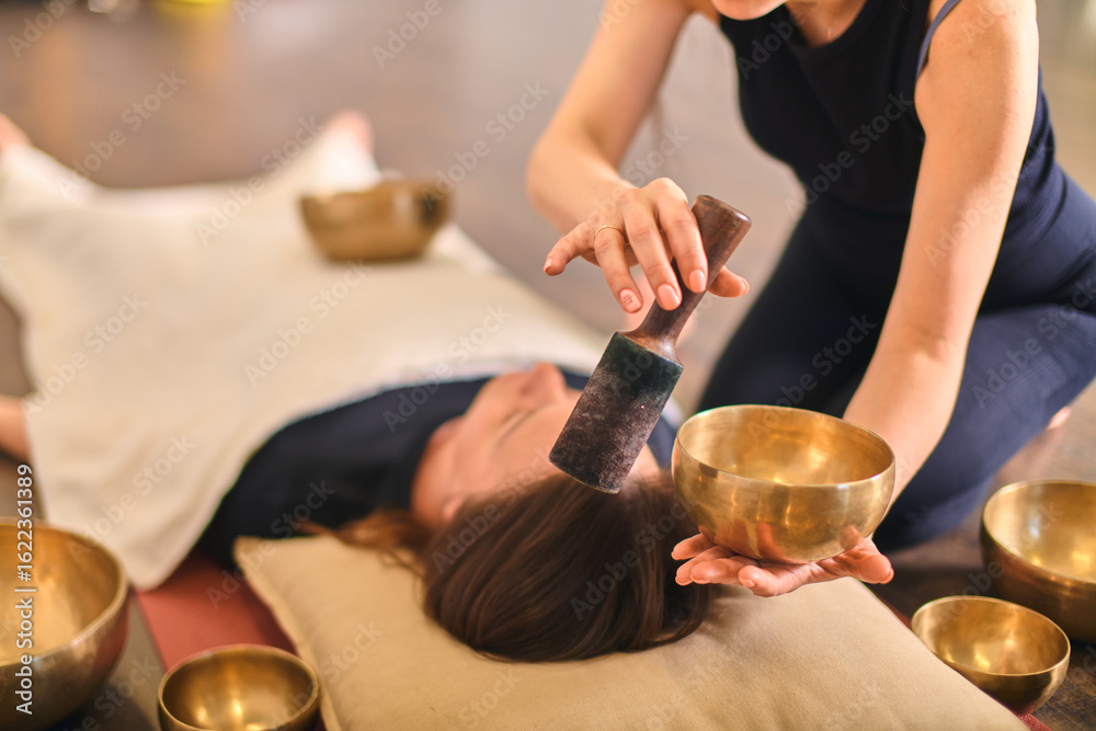 Fototapeta premium Woman using singing bowl for sound therapy over lying person. Warm lighting enhances calming atmosphere. Bowls placed strategically around for holistic relaxation