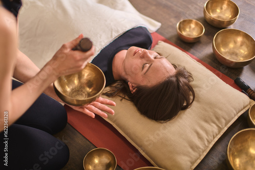Young woman at peace during sound therapy session. Therapist uses singing bowls for relaxation. Warm lighting on wooden floor enhances tranquil atmosphere