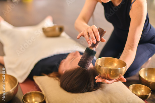 Woman using singing bowl for sound therapy over lying person. Warm lighting enhances calming atmosphere. Bowls placed strategically around for holistic relaxation