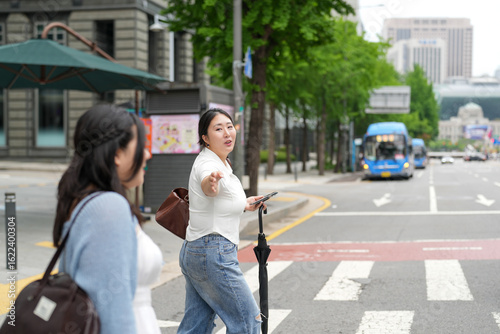 Canvas Print Two Korean women in their twenties in casual clothes walk across a crosswalk on a boulevard, talking together and smiling, carrying bags beneath bright spring light