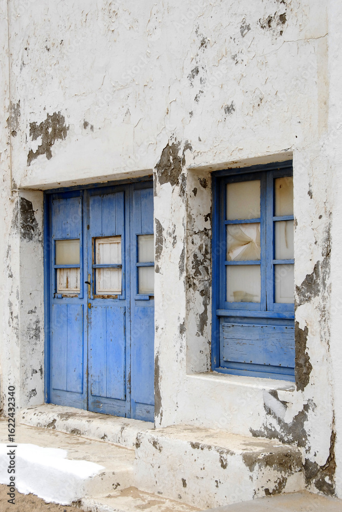 Fototapeta premium A faded blue door and matching window stand on a crumbling whitewashed wall. The aged textures tell a quiet story of time, abandonment, and Mediterranean charm.