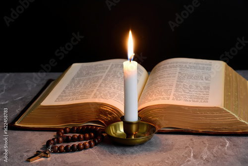 A lit candle in a brass holder sits on an open bible with prayer beads in front