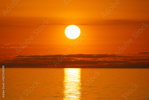 Panorama of a bright orange sunset with the sun fully visible over the sea and long clouds on the horizon