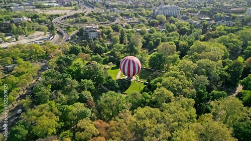 Arc tracking aerial drone video of the FlyBalloon in Budapest’s City Park, centered and framed by trees, with Budapest Környéki Törvényszék, tram stop, and main crossroads in the background.
