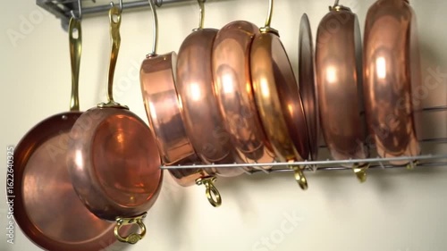 Rotating Wall Mounted Copper Cookware Rack Displaying Assorted Pots and Pans with Brass Handles in Bright Kitchen with Cream Wall and Partial White Cabinet