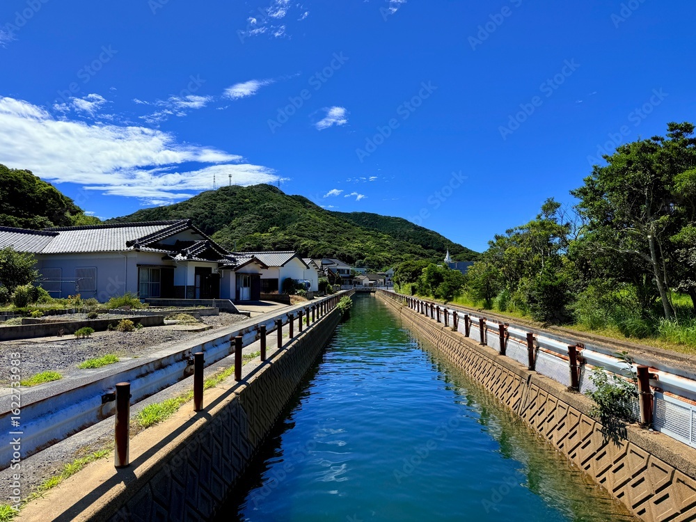 Fototapeta premium Seaside scenery of Naru Island, Goto Islands, Nagasaki, Japan – traditional houses, canal, and crystal-clear coastal waters surrounded by lush green hills and summer skies.
