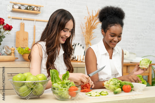 Black and white teenage friends happily and joyfully together to making a vegetable salad in a white kitchen. They enjoy healthy food.