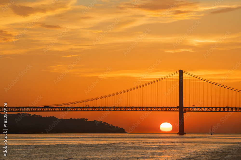 Fototapeta premium Sunset view of the 25 de Abril Bridge in Lisbon over the Tagus River. Perfect for postcards magnets travel guides magazines websites and tourism materials.