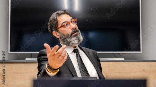 Картината върху платно A middle-aged, dark-haired, bearded businessman in a suit is sitting at a confer