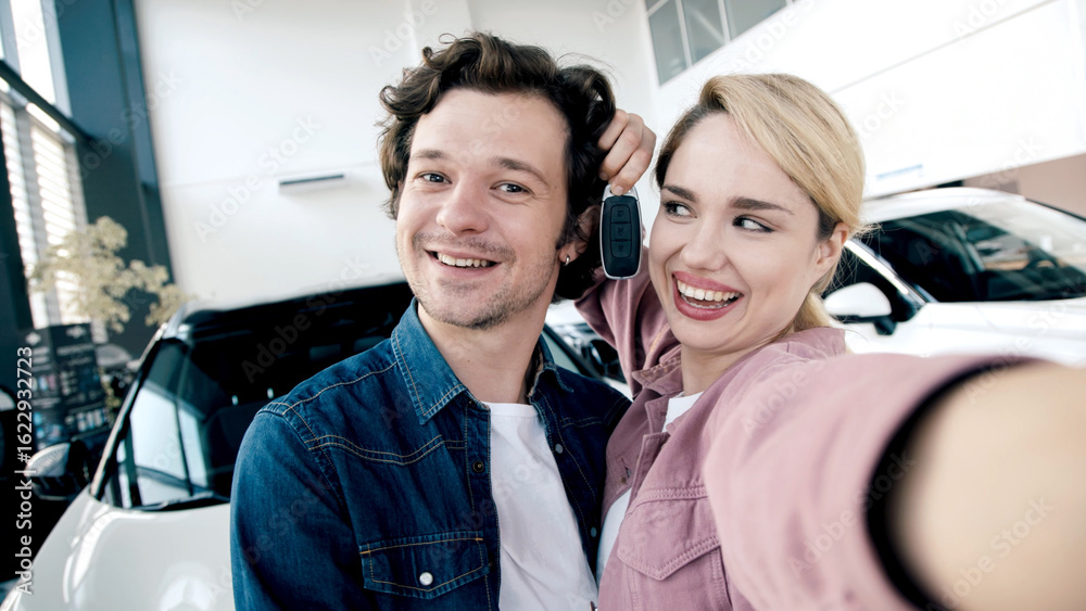 Fototapeta premium Happy couple taking selfie in car showroom, showcasing excitement after purchasing new vehicle, surrounded by modern cars and bright atmosphere