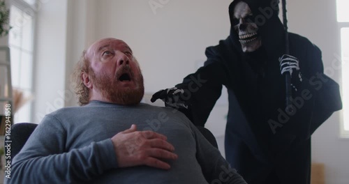 Portrait of senior shocked scared dying old man in casual clothes sitting on chair at home with person in grim reaper death costume holding scythe standing behind him in living room. 4k video.