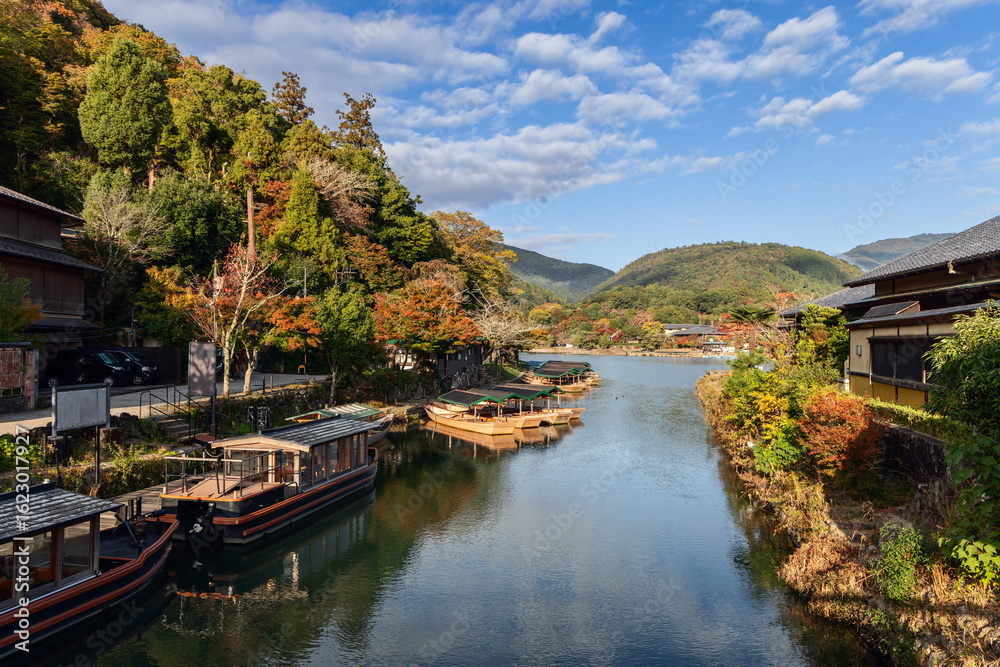 Fototapeta premium Wooden boats line the calm river in Arashiyama, Kyoto, Japan, with autumn trees on both banks and forested mountains in background under clear sky in early light