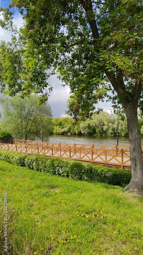 Wooden bridge on bank of navigable river. Well-maintained embankment in city park. Spring or summer landscape. Place to walk and relax after work. Cozy atmosphere of nature park. Vertical photo Wooden bridge on bank of navigable river. Well-maintained embankment in city park. Spring or summer landscape. Place to walk and relax after work. Cozy atmosphere of nature park. Vertical photo