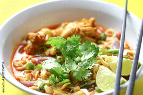 A bowl of spicy noodle soup with chicken, garnished with fresh cilantro, lime wedges, and chopped vegetables, served with black chopsticks. The vibrant colors and presentation are visually appealing.