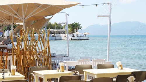 Wallpaper Mural Coastal restaurant in Aegina, Greece with tables under a light umbrella, dry branch decor, and calm sea in the background. Distant café, palm trees, and mountains visible. Sunny and relaxing Torontodigital.ca